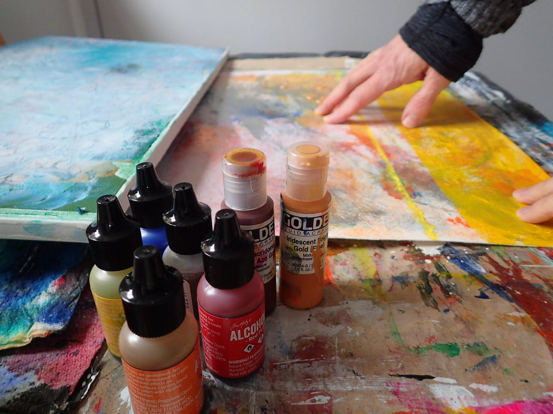 Kimberli Werner's hand touching an abstract painting, with other paintings scattered around the table, and seven paint bottles in the foreground.